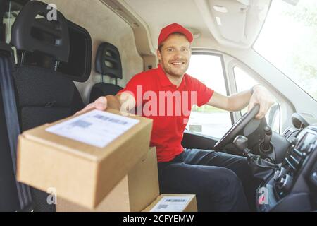 giovane sorridente addetto al servizio di consegna seduto in furgone e consegna scatole Foto Stock