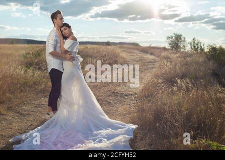 giovane coppia di sposa e sposo in abito da sposa, vestito bianco, sul campo, sullo sfondo del cielo blu nuvoloso. Tiro al matrimonio, sposa An Foto Stock