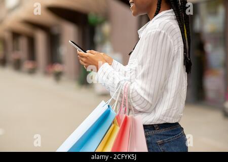 Incontro vicino al negozio per lo shopping. Sorridente donna afro-americana blogger digitando su smartphone e tenendo borse Foto Stock