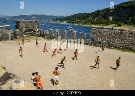 Gruppo di teenager che giocano a calcio nella parvenza della Chiesa di San Pietro con l'Isola Palmaria sullo sfondo, Porto Venere, la Spezia, Italia Foto Stock