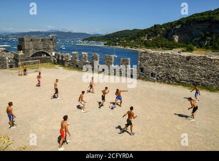 Gruppo di teenager che giocano a calcio nella parvenza della Chiesa di San Pietro con l'Isola Palmaria sullo sfondo, Porto Venere, la Spezia, Italia Foto Stock