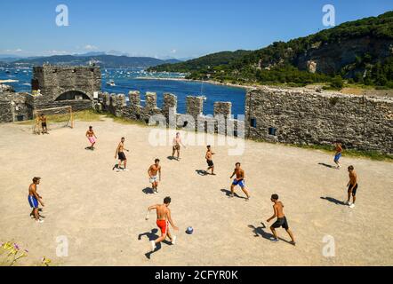 Gruppo di teenager che giocano a calcio nella parvenza della Chiesa di San Pietro con l'Isola Palmaria sullo sfondo, Porto Venere, la Spezia, Italia Foto Stock