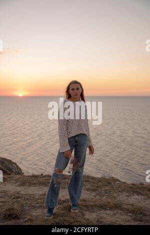 Una ragazza bionda in jeans trapelati si pone sullo sfondo del tramonto . Foto Stock