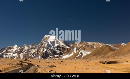 Immagine di messa a fuoco selettiva di una lunga strada solitaria con curve sull'altopiano tibetano con montagne e vette ricoperte di neve e cielo blu chiaro di fronte e tibe Foto Stock