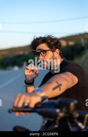 Giovane uomo alla moda seduto e fumando su una moto classica al tramonto. Foto Stock