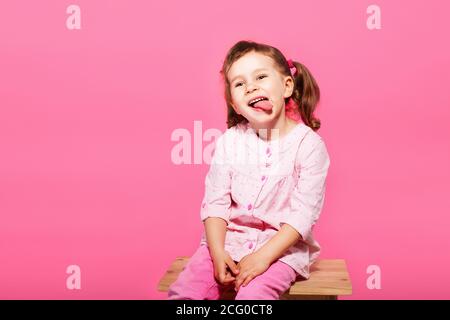 Bambino che gioca la scimmia. Bambina che indossa abiti rosa su sfondo rosa. Ragazza che mostra la lingua e divertirsi. Felice infanzia Foto Stock