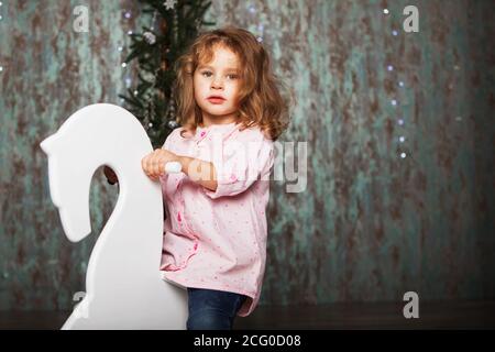 Ritratto di una bella bambina. Ritratto di bambina carina tra le decorazioni di Natale. Ragazza che cavalca un cervo a dondolo. Sedia dondolo cervi per bambini ri Foto Stock