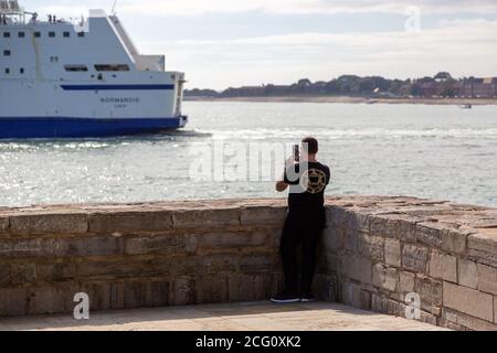 Un uomo che usa il suo telefono cellulare o cellulare per scatta una foto di un traghetto che passa accanto al mare Foto Stock