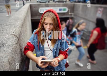 La donna in una felpa con cappuccio rossa legge i messaggi sullo schermo dello smartphone vicino all'ingresso della metropolitana. Foto Stock