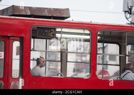 BELGRADO, SERBIA - 14 agosto 2020: Giovane uomo con cuffie alla finestra di un tram che indossa una maschera respiratoria viso durante i mezzi pubblici Foto Stock