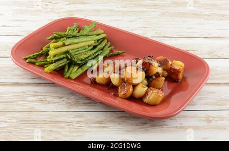 Capesante con salsa di pancetta allo zucchero di canna e asparagi cosparsi di aglio in polvere su piatto di portata rosso Foto Stock