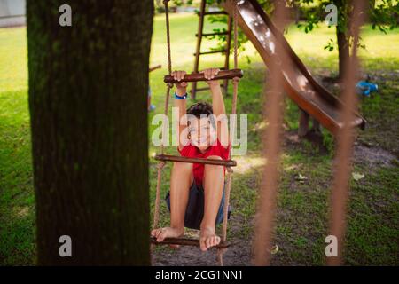 Un bel ragazzo caucasico su una scala di legno per bambini nel parco. Un giorno in natura Foto Stock