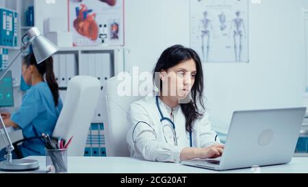 Giovane donna dottore in gabinetto medico privato digitando sul computer portatile, mentre l'infermiera in lavoro in background. Sistema sanitario medico operaio professionista in ospedale e ricerca sanitaria Foto Stock