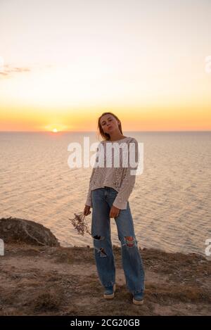 Una ragazza bionda in jeans trapelati si pone sullo sfondo del tramonto . Foto Stock