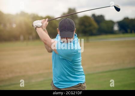 Uomo che gioca a golf oscillando alla palla come gioca il suo scatto utilizzando un driver visto da dietro guardando verso il basso il fairway in un sano stile di vita attivo co Foto Stock