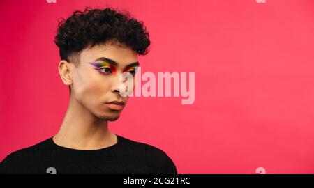 Primo piano di un uomo fluido di genere con il trucco dell'occhio arcobaleno guardando via su sfondo rosso. Uomo transgender con bandiera di orgoglio occhio ombra. Foto Stock