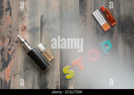 Sigarette nere di e-sigaretta, e-liquido dolce, sigarette di tabacco con accendino e parola di lettere di legno su sfondo grigio legno coperto di fumo. Vista dall'alto. Foto Stock