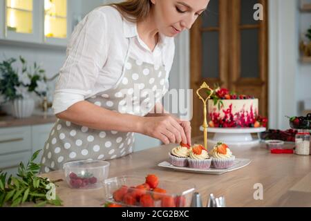 donna in grembiule adorna le cupcake con le fragole, mentre lo chef della pasticceria in cucina prepara dessert, torte e muffin. Cucinare a casa Foto Stock