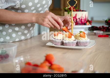 Donna in grembiule adorna cupcake con fragole, pasticciere in cucina produce dessert, torte e muffin. Cucinare a casa. Foto Stock