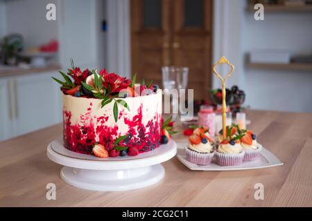 Grande bella torta di velluto rosso, con fiori e bacche in cima. Deliziosi muffin dolci con crema, decorati con fragole e mirtilli. Foto Stock