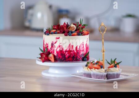 Grande bella torta di velluto rosso, con fiori e bacche in cima. Deliziosi muffin dolci con crema, decorati con fragole e mirtilli. Foto Stock