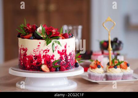 Grande bella torta di velluto rosso, con fiori e bacche in cima. Deliziosi muffin dolci con crema, decorati con fragole e mirtilli. Foto Stock