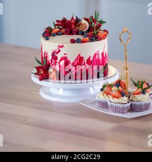 Grande bella torta di velluto rosso, con fiori e bacche in cima. Deliziosi muffin dolci con crema, decorati con fragole e mirtilli. Foto Stock