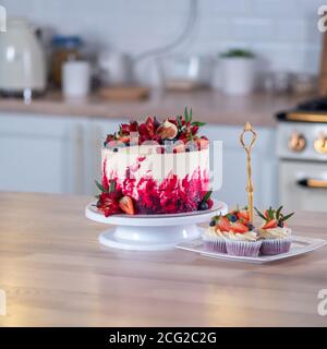 Grande bella torta di velluto rosso, con fiori e bacche in cima. Deliziosi muffin dolci con crema, decorati con fragole e mirtilli. Foto Stock