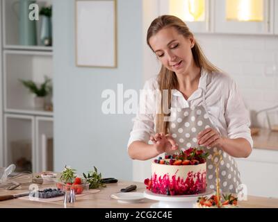 Cottura e decorazione di torta con crema. Giovane donna pasticciera in cucina decorando la torta di velluto rosso. Donna felice in grembiule in cucina. Foto Stock