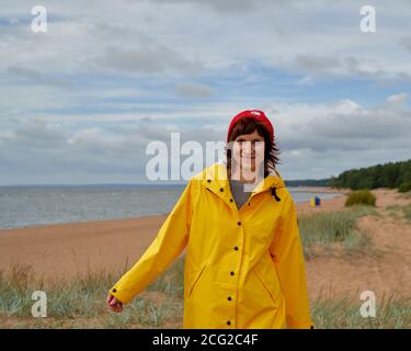 Vita ritratto di donna matura in impermeabile giallo e cappello rosso camminando lungo la spiaggia e guardando la macchina fotografica, sorridendo. Autunno passeggiata nella natura in abiti luminosi. Bilanciamento femminile sulla spiaggia Foto Stock