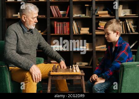 Il nonno e il nipote senior giocano a scacchi seduti alla scrivania in un accogliente soggiorno. Foto Stock