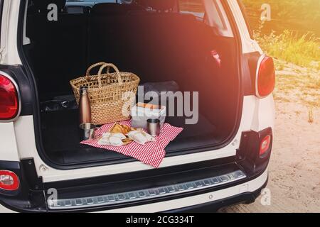 Pacchetto picnic e cestino con croissant e panini si trovano nel bagagliaio dell'auto da berlina. Weekend all'aperto, viaggi locali. Foto dai toni soleggiati Foto Stock