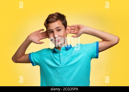 Il concetto di adolescenza ed emozioni negli adolescenti. Un ragazzo adolescente in una t-shirt blu, allegro tirando le orecchie con le mani. Sfondo giallo Foto Stock