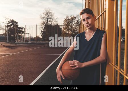 Sport e basket. Un giovane adolescente in una tuta nera posa con un basket, in piedi alla recinzione. Spazio di copia. Foto Stock