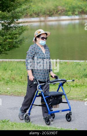 Una donna asiatica americana più anziana che indossa una maschera chirurgica e spinge il suo camminatore, cammina intorno a un lago in un parco Flushing, Queens, New York. Foto Stock