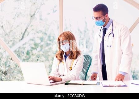 Foto di dottore di sesso femminile seduto alla scrivania dietro il suo computer e consultandosi con un medico di sesso maschile nella stanza del medico. Medici che indossano maschere per la prevenzione Foto Stock