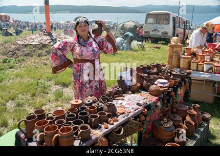 Una donna di vendita con una caraffa di terracotta in mano si trova dietro un bancone con piatti al festival di musica e artigianato di Caratag. Regione di Krasnoyarsk. Foto Stock
