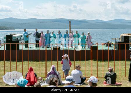 Gli spettatori guardano l'ingresso delle donne sul palco, sullo sfondo del lago, al festival musicale Caratag. Regione di Krasnoyarsk. Russia. Foto Stock