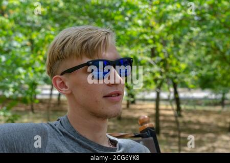 Ritratto di giovane uomo elegante che indossa occhiali da sole. Bell'uomo giovane in occhiali da sole eleganti con un'elegante acconciatura poggiata sulla panca del parco. Li attivo Foto Stock