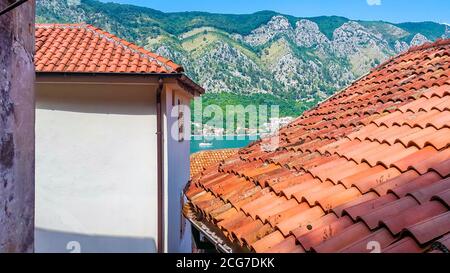 Vista dall'alto dei tetti in tegole rosse sulla città vecchia di Cattaro (Montenegro), le montagne e le acque blu della baia di Boka Kotor con barca a vela dietro. Foto Stock