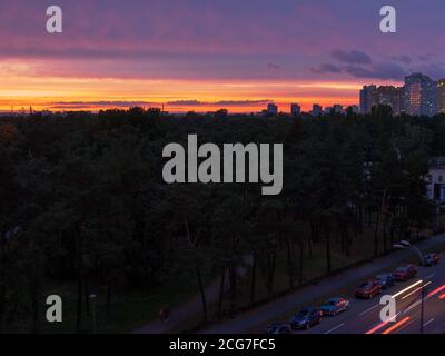 Sorprendente viola, viola, rosso e giallo tramonto su Kiev città con edifici con luce in finestre e strada con auto in movimento con fari. Foto Stock