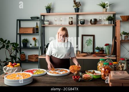 Donna matura bionda in piedi al bancone in cucina accogliente e. stendendo le fette di frutta su un vassoio di plastica mentre si fanno spuntini sani Foto Stock