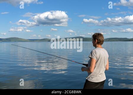 Una donna pescatrice cattura il pesce per un'esca da un lago blu sullo sfondo di una costa montana. Scatto luminoso. Foto Stock