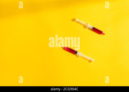 Due siringhe monouso in plastica con sangue rosso o vaccino su sfondo giallo con spazio di copia ombreggiato. Foto Stock