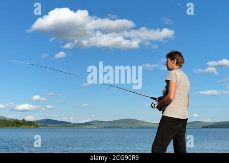 Donna pescatrice cattura un pesce da un lago blu contro un cielo blu con le nuvole. Foto Stock