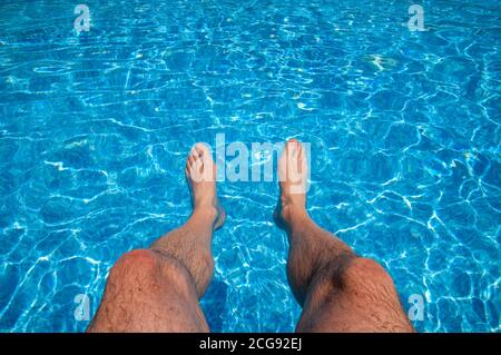Coppia maschile di piedi e gambe inferiori penzolanti dal bordo di una piscina con acqua turchese increspata. Foto Stock