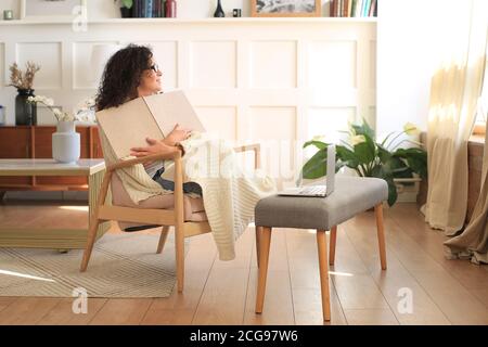 Bella donna con capelli ricci coperto di una plaid legge a. prenota in una sedia Foto Stock