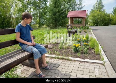 Un ragazzo pensivo di 11 anni si siede su una panchina di legno nel parco in una giornata estiva di sole. Foto Stock