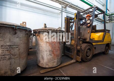 Magazzino di impianti di metallurgia del titanio. Operaio asiatico su carrello elettrico giallo con secchio di stoccaggio con spugna di titanio. Foto Stock