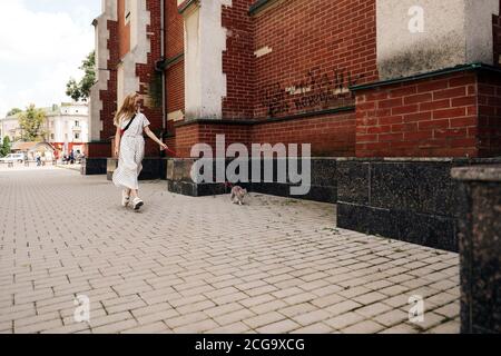 Una giovane donna cammina con un gatto scozzese dritto sulla strada a guinzaglio in città. Ragazza bionda vestita con un lungo abito bianco Foto Stock